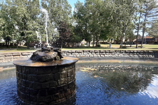 A photo of a brick-lined pond. In the centre of the pond is a large, brick mound with a water fountain. The fountain is shooting water up into the air.