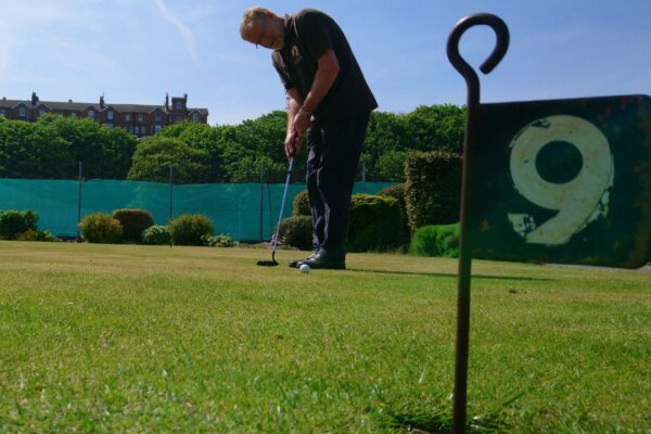 A photo of a person putting a golf ball. In the foreground a metal flag with the number 9 is in a hole