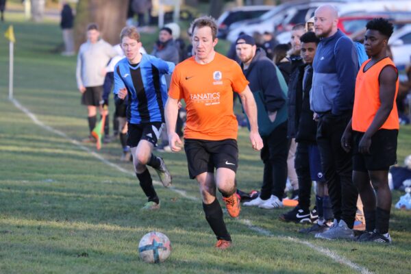 The photo shows two people playing football. A group of people watch from the sidelines