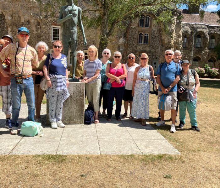 A group of people standing smiling at the camera. They are stood outside near a statue of a person in front of a country house