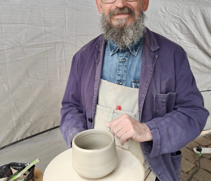 A person smiles at the camera. They are sat behind a potter's wheel