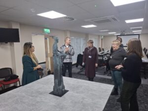 Five people stand gathered around a table talking. On the table is a statue of a child on a rock.